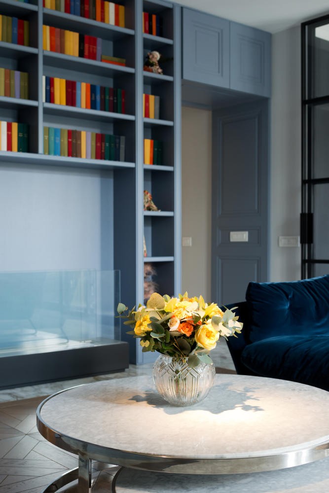 0-American-style-interior-living-room-lounge-bio-fireplace-home-library-book-shelves-blue-velvet-sofa-wall-panelling-metal-round-coffee-table-flowers-vase
