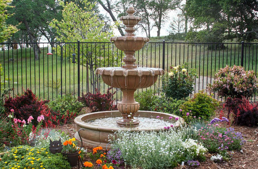 Stone fountain in a garden surrounded by greenery and a metal fence