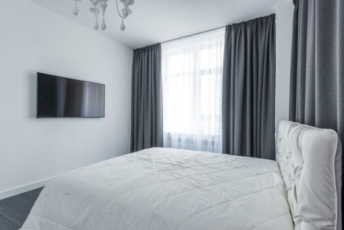 pexels-7587777 A modern bedroom featuring minimalist decor with a flat-screen TV and elegant curtains.