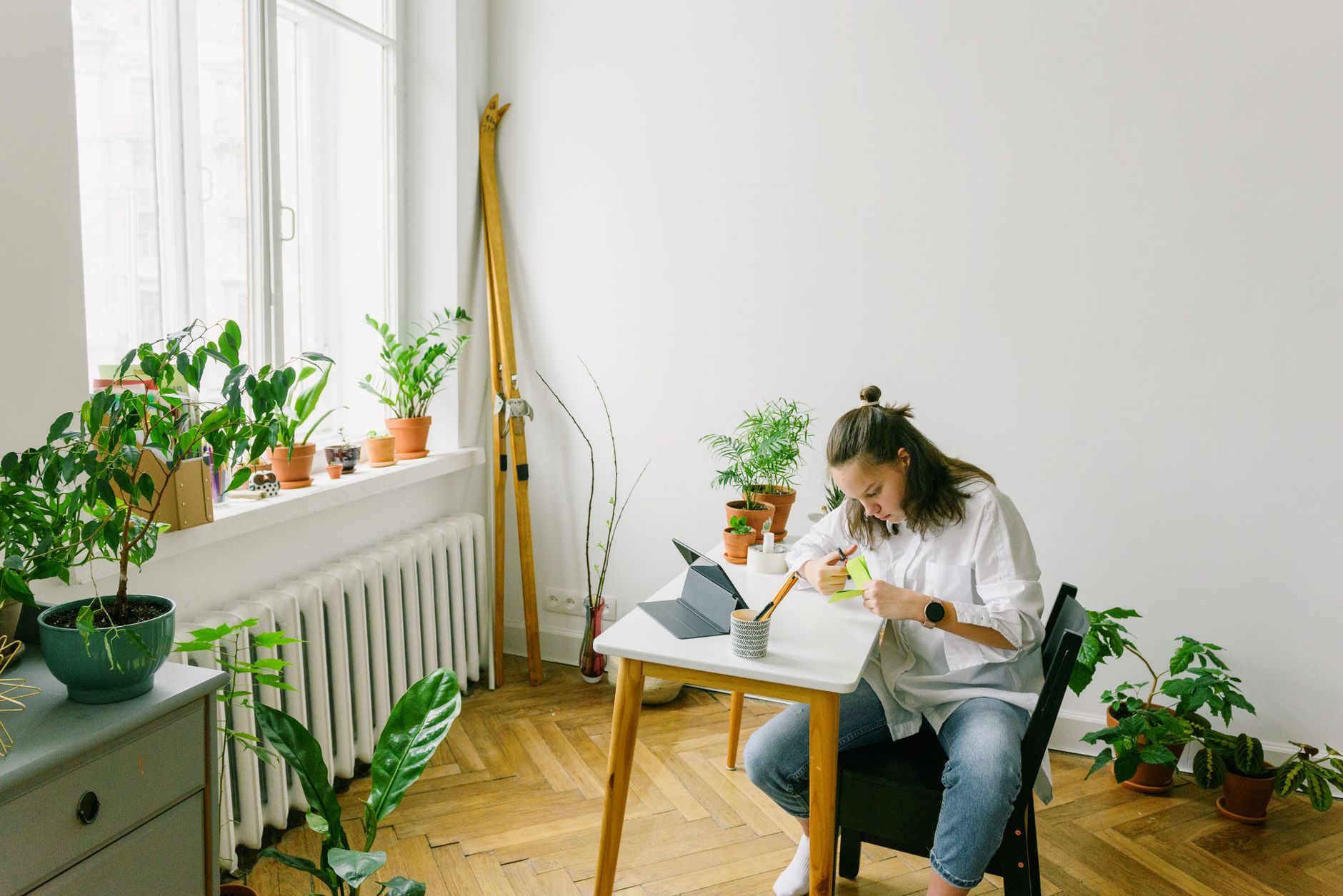 A woman sits at a table in a bright room filled with plants, writing notes with focus.