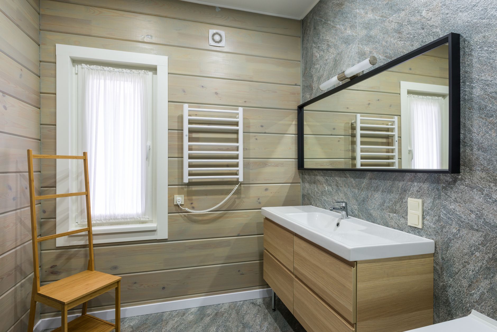 Light bathroom interior with sink under mirror hanging near paneled wall with window covered with curtain