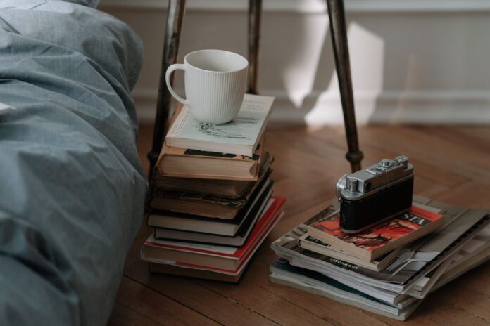 A cozy bedroom corner featuring vintage camera, piles of books, and a white cup, creating an inviting atmosphere.