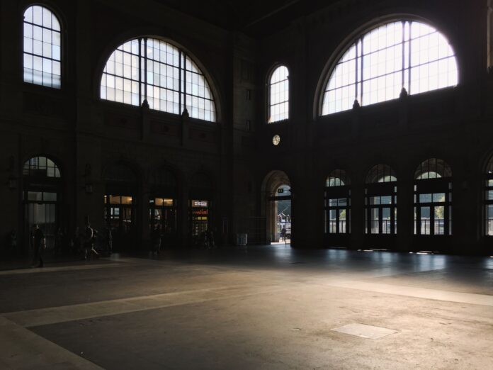 Sunlit interior of a spacious building hall with large arched windows casting shadows.