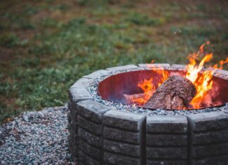 8 Backyard Fire Pit Ideas for Cozy Fall Evenings Outdoors Old dry flaming piece of log in big stone round fire bowl on ground covered with pebbles on lawn in daylight