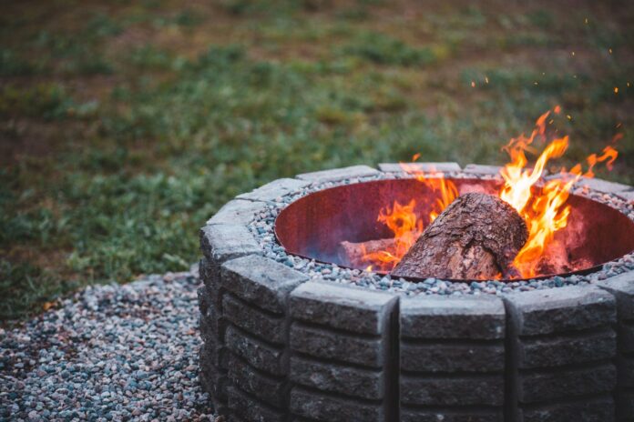 Old dry flaming piece of log in big stone round fire bowl on ground covered with pebbles on lawn in daylight