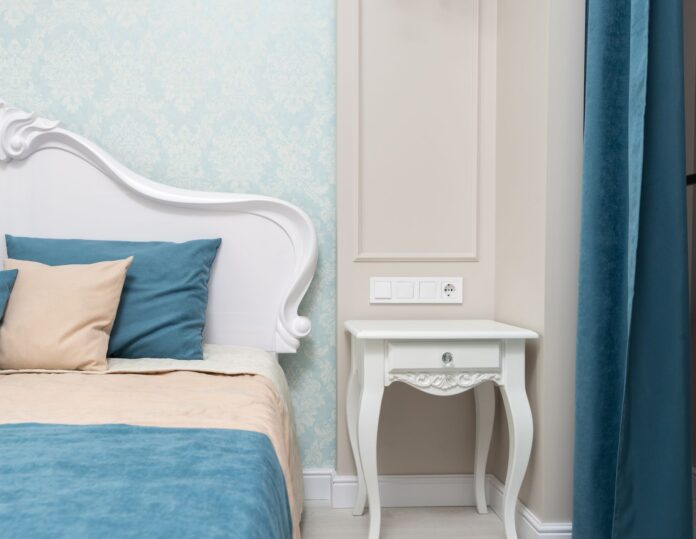 Comfortable bed with colorful bedclothes and cushions placed at wall with wallpaper near bedside table and curtain in bedroom with classic interior