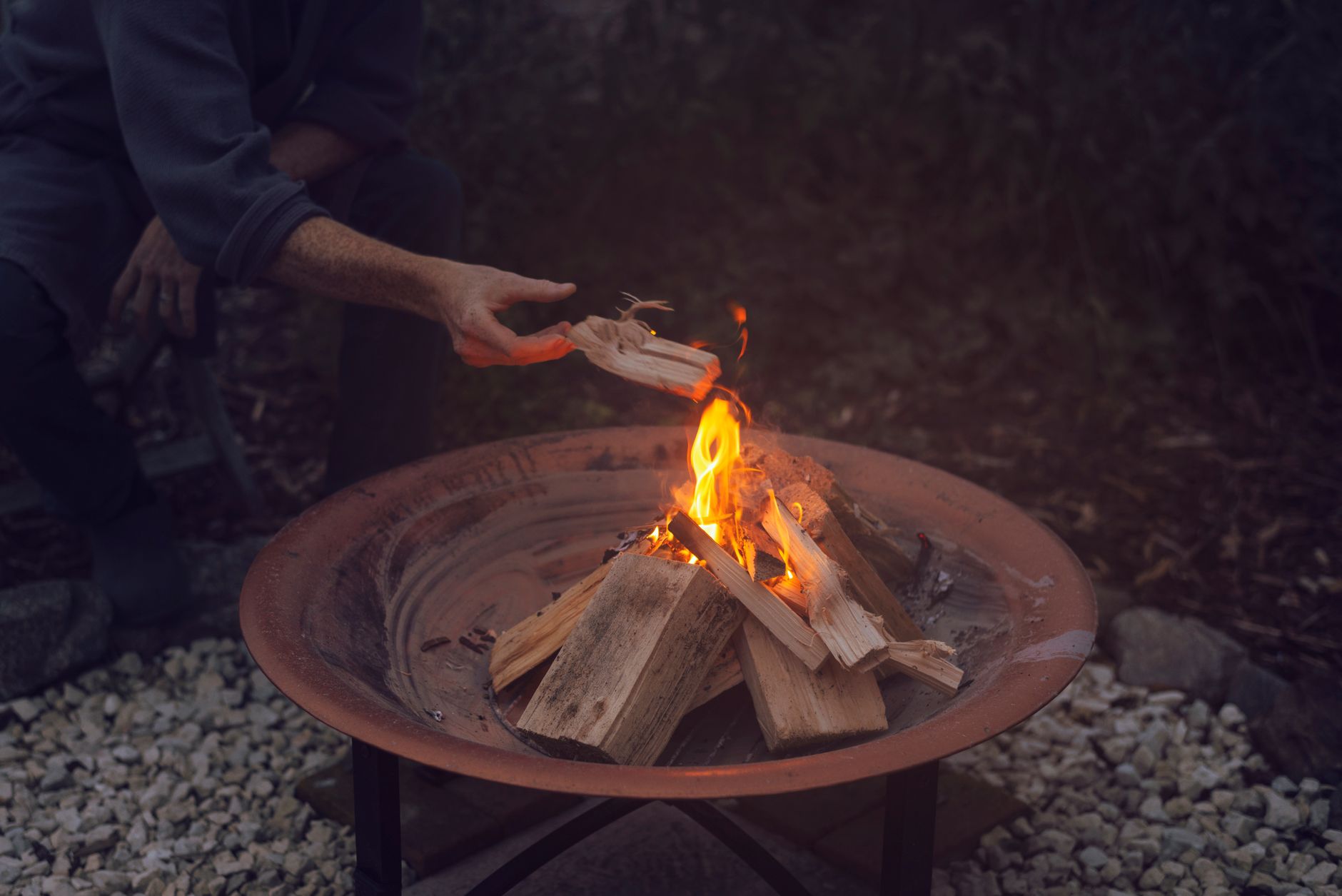 A man adding wood to an outdoor campfire pit on a tranquil day.